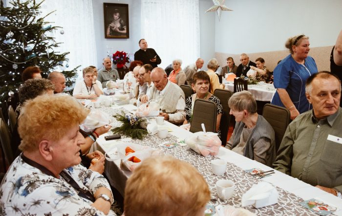 mieszkańcy siedzący przy stolach oraz ksiądz który składa im życzenia